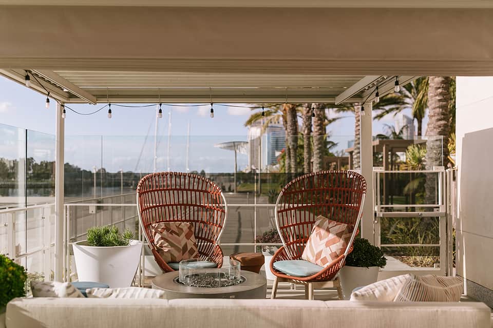 Modern rooftop lounge with rattan chairs, fire pit table, glass railings, and palm trees
