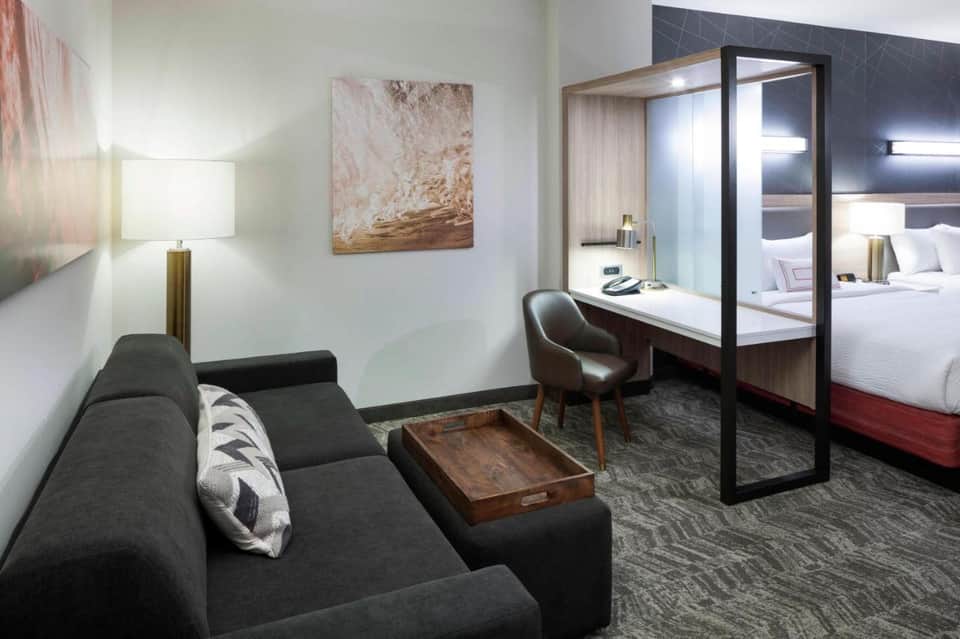 Hotel suite with living area, modern desk, open bathroom with red accents and glass partition