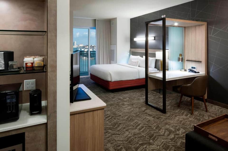 Modern hotel room with king bed, glass-enclosed bathroom, work area, and balcony
