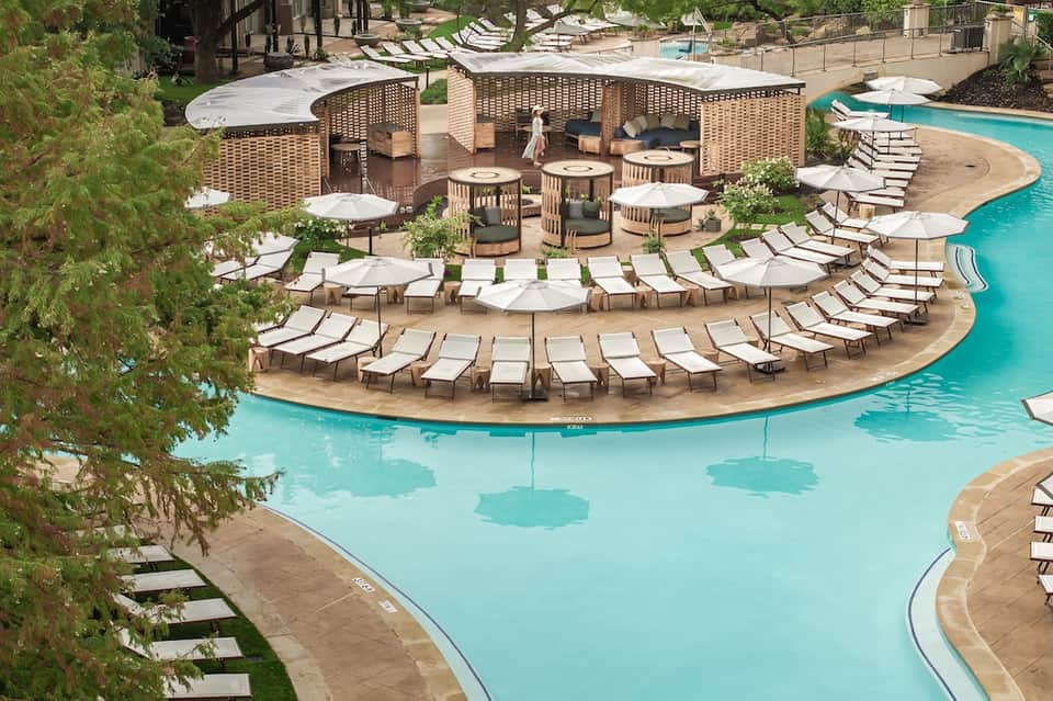 Curved resort pool with lounge chairs, white umbrellas, and wooden cabanas among tropical landscaping
