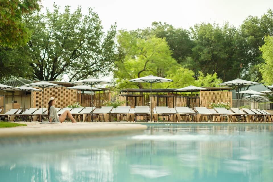 Luxury resort pool area with wooden loungers, white umbrellas, and lush tree canopy backdrop