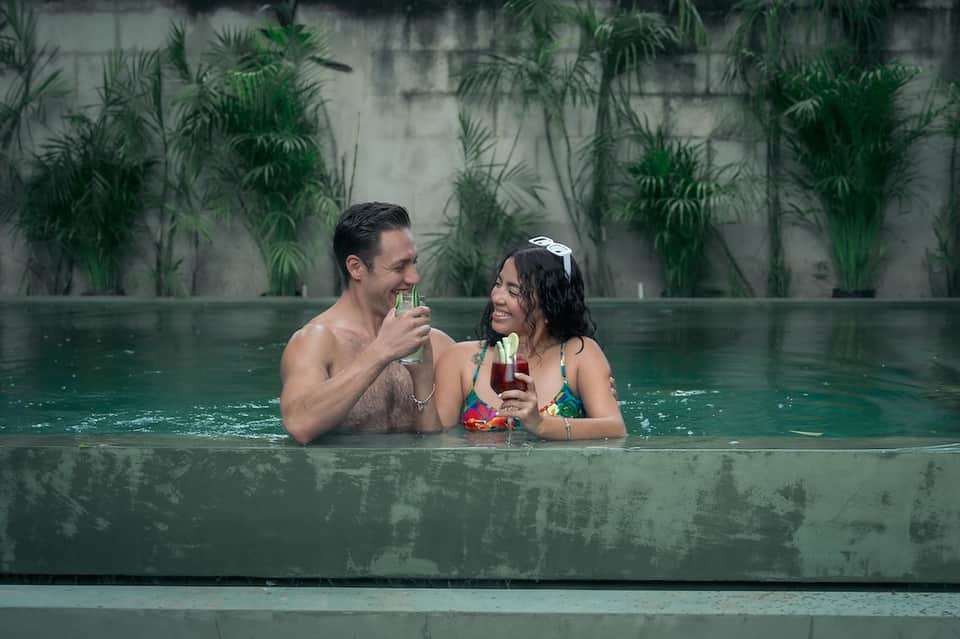 Couple enjoying cocktails in an infinity pool with tropical foliage backdrop and concrete walls