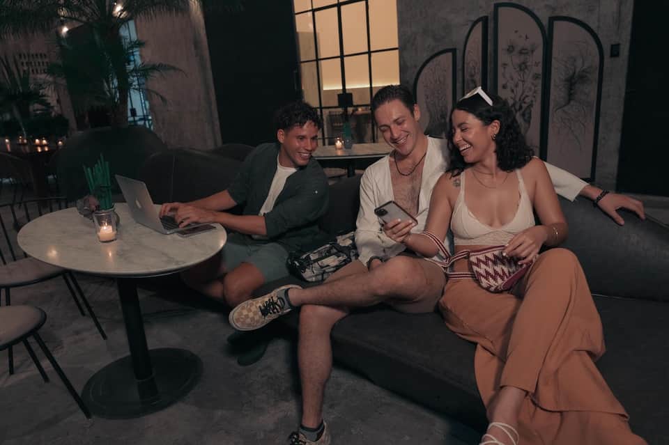Three guests relaxing on outdoor sofa at night with laptop and cocktails in modern resort setting