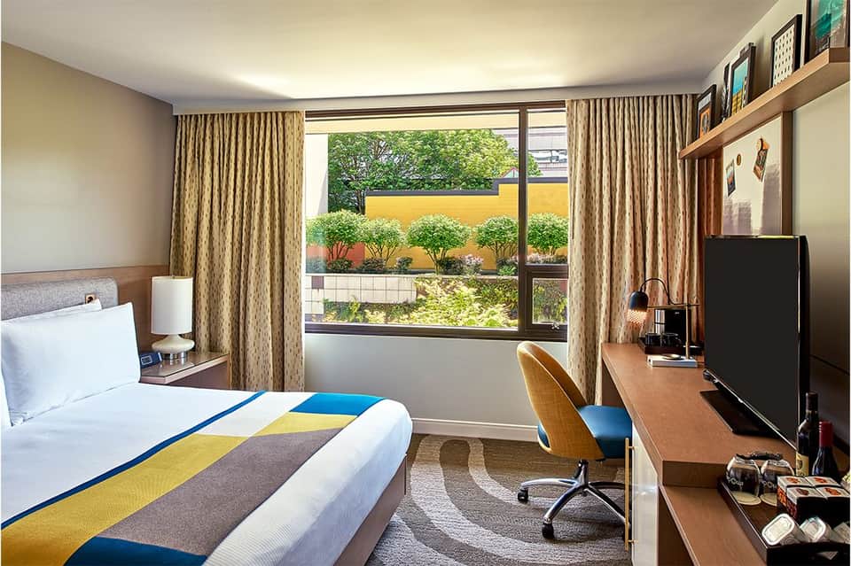 Contemporary hotel bedroom with colorful bedding, work desk, window overlooking garden with yellow accent wall