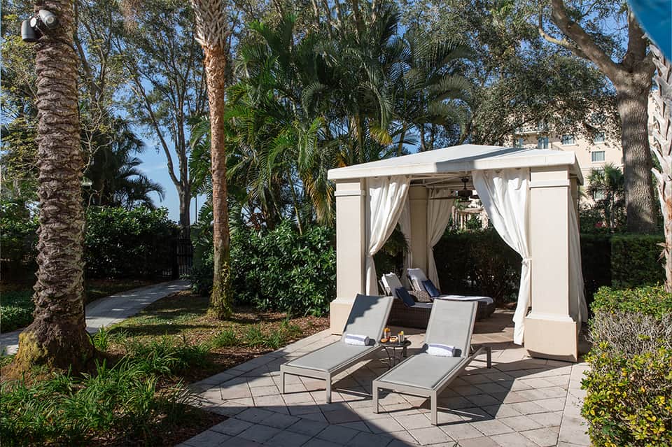 Outdoor cabana with white curtains, lounge chairs, surrounded by palm trees and landscaping