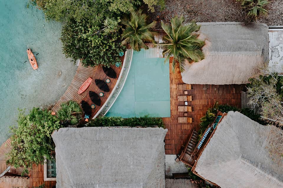 Aerial view of resort pool, wooden deck, palm trees, and turquoise water surrounded by buildings