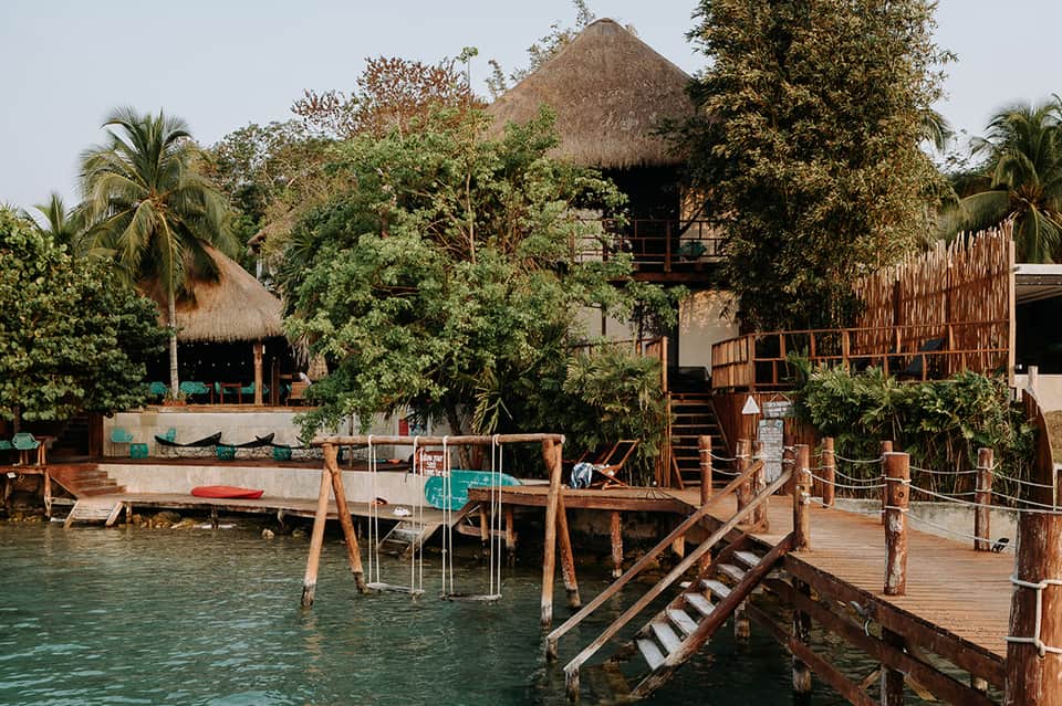 Tropical waterfront resort with thatched-roof buildings, wooden docks, and lush vegetation