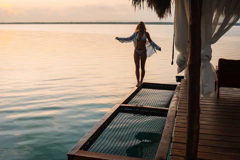 Woman in bikini standing on wooden dock at sunset, ocean view with palm fronds overhead