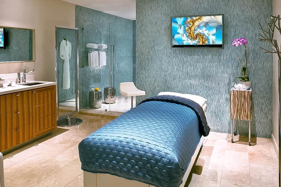 Spa treatment room with teal textured walls, massage bed, modern bathroom, and relaxation amenities