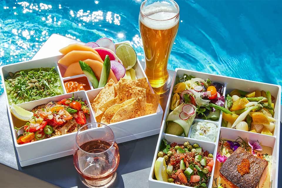 Colorful meal boxes with fresh vegetables, ceviche, and beer displayed by sparkling blue pool
