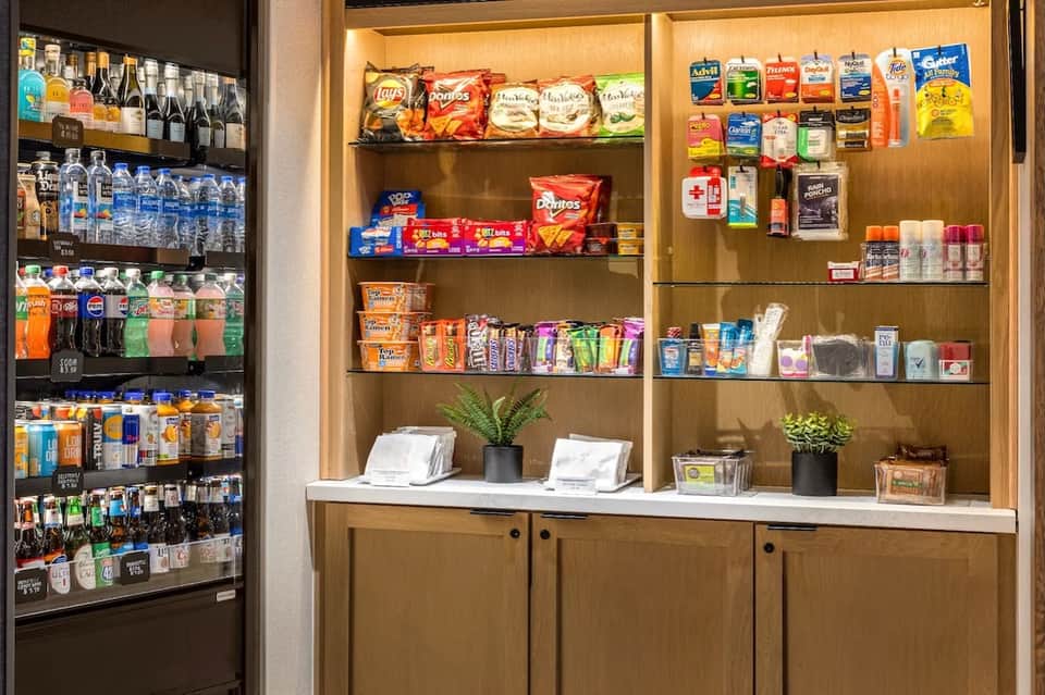 Hotel beverage station with refrigerated drink display, wooden shelves stocked with snacks and toiletries