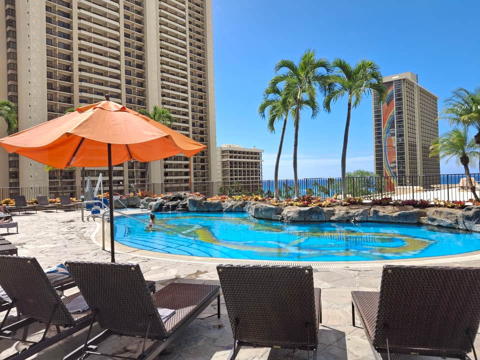 Beachfront resort pool with turquoise water, orange umbrella, palm trees, high-rise buildings, and ocean views