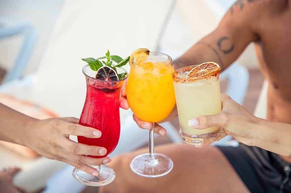 Hands holding three colorful cocktails with citrus garnishes and ice at a resort pool party