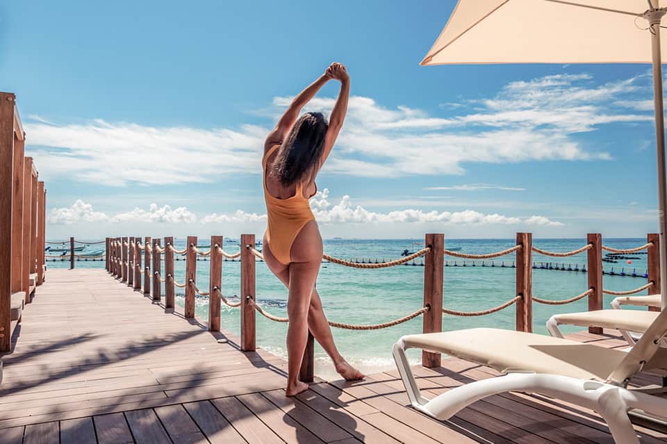 Woman in tan swimsuit stretching on wooden beachfront deck with turquoise ocean, lounge chairs, and umbrella in background
