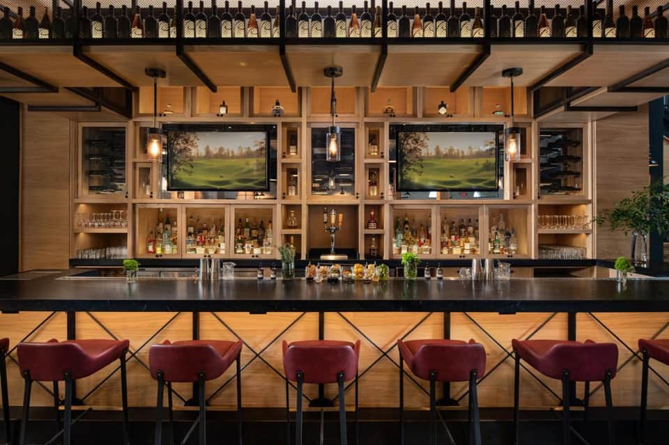 Upscale bar interior with dual TV screens, liquor shelves, and burgundy bar seating