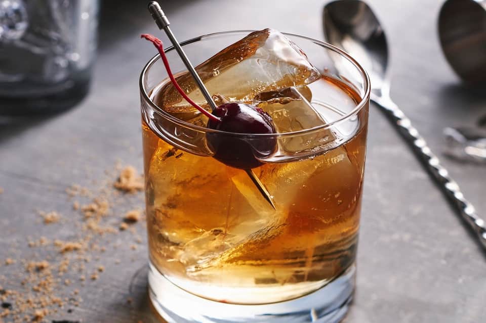 Cocktail with amber liquid, ice, and cherry garnish with red stirrer on gray surface