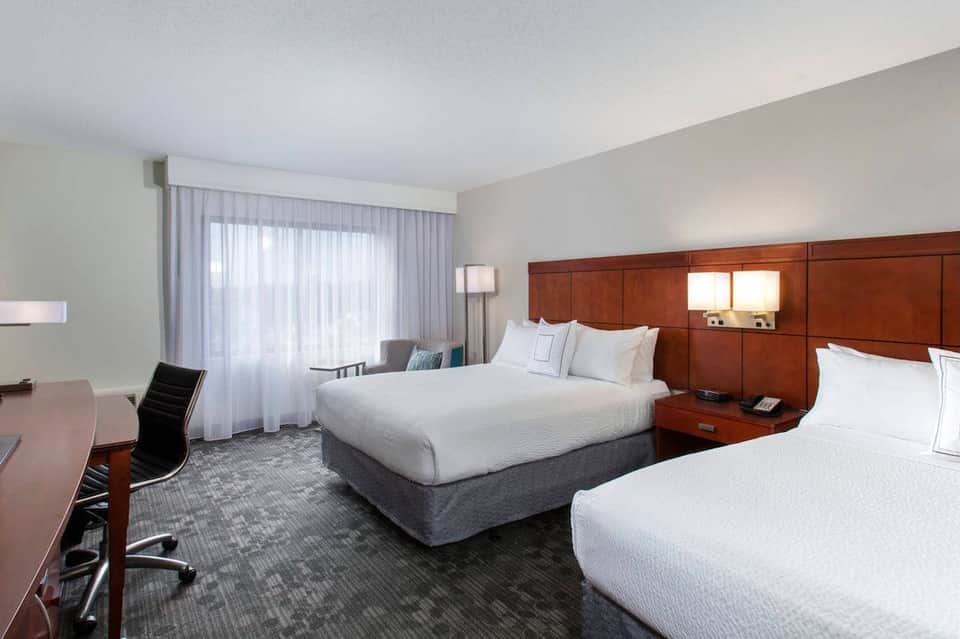 Hotel double queen bedroom with wood headboard, desk, and large windows with curtains