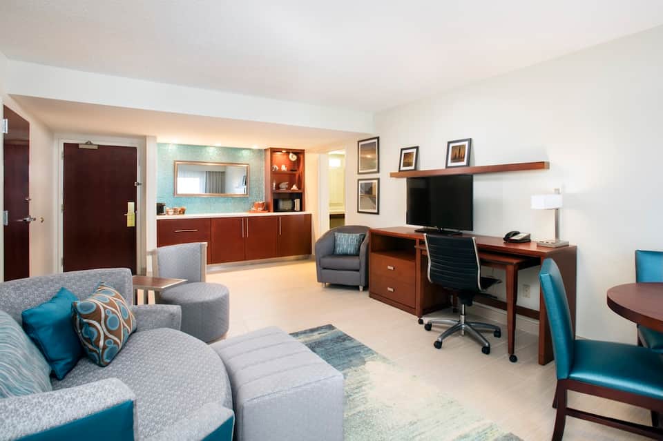 Spacious hotel suite with living area, desk, kitchenette, and wood furnishings in neutral tones