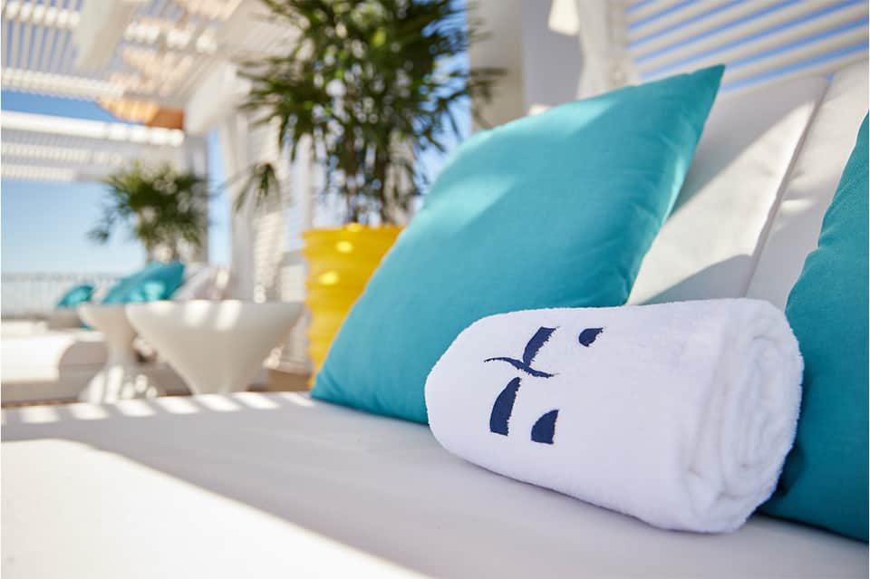 Close-up of turquoise pillow on white lounge chair with striped umbrella and plants in background