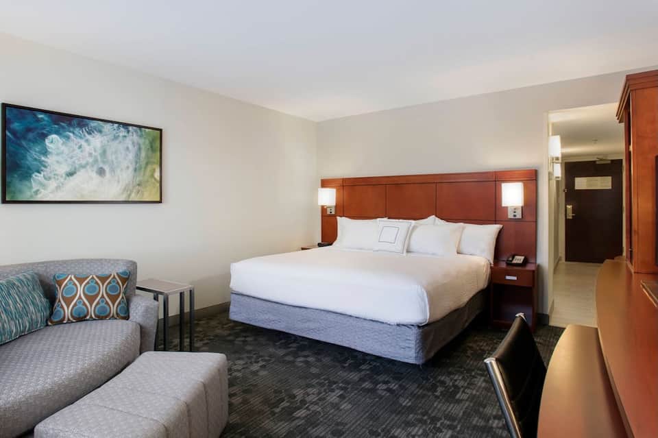 Hotel bedroom with queen bed, wood headboard, seating area, and flat-screen TV on wall
