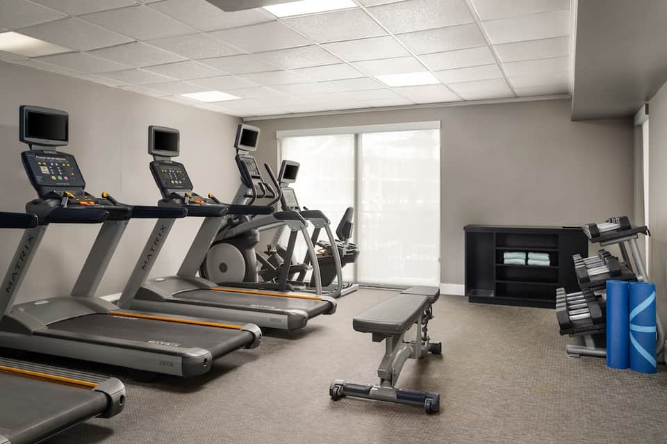 Hotel gym with multiple treadmills, elliptical machines, and weight benches near sliding glass doors