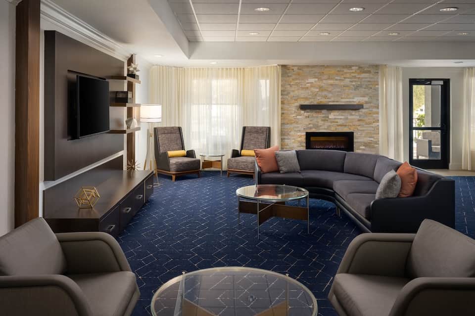 Modern lobby lounge with curved gray sofa, fireplace, TV, blue patterned carpet, and stone wall