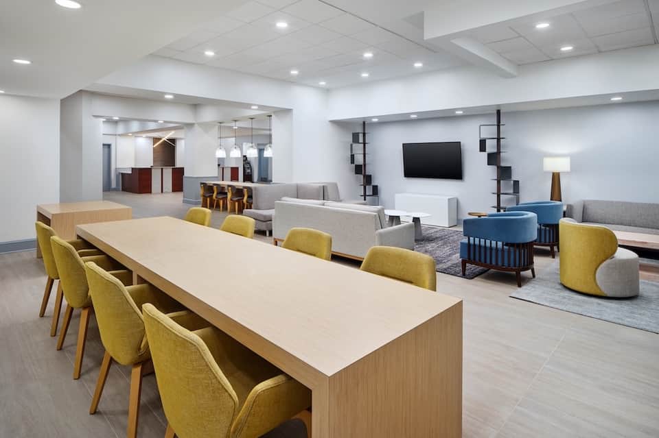 Modern hotel gathering space with dining table, yellow chairs, lounge seating, and reception desk