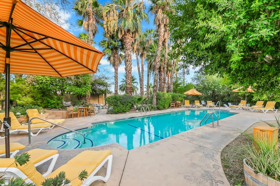 Resort pool area with turquoise water, yellow lounge chairs, orange umbrellas, and palm trees on sunny day