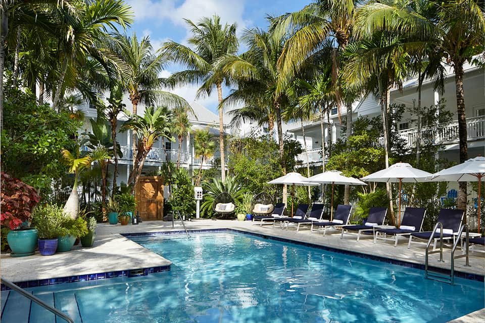 Resort pool surrounded by palm trees, white umbrellas, and colonial-style buildings