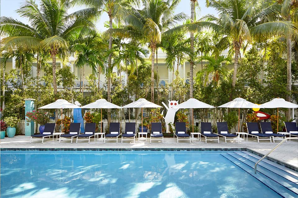 Resort pool with white umbrellas, lounge chairs, palm trees, and colonial-style building in background