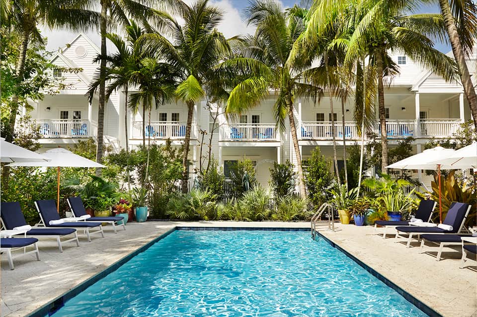 Resort courtyard with rectangular pool, white colonial buildings, palm trees, and lounge seating