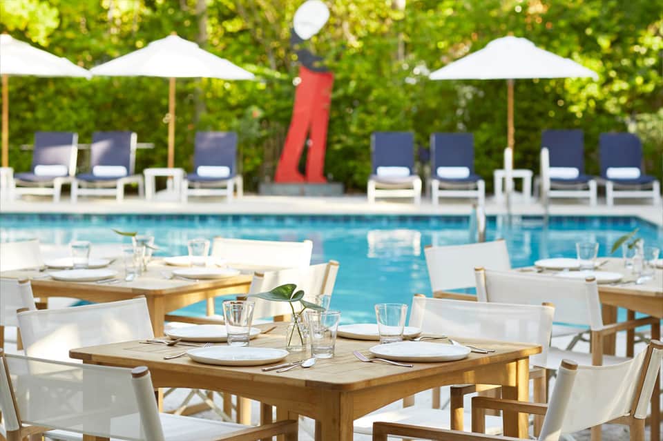 Poolside dining area with wooden tables, white chairs, umbrellas, and lounge seating