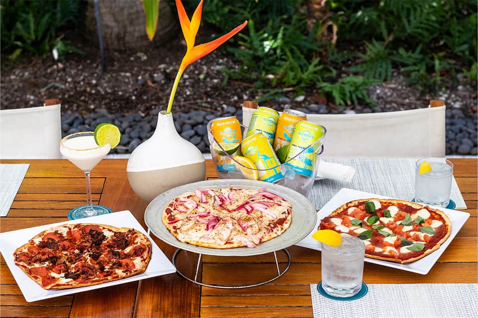 Outdoor dining table with three colorful pizzas, tropical cocktails, and bird of paradise flower centerpiece