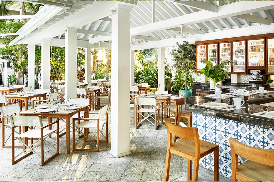 Covered dining patio with white columns, wooden tables, blue accents, and tropical plants