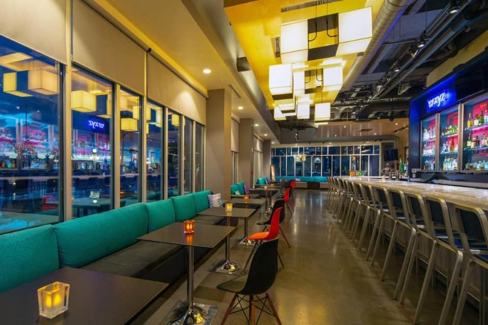 Contemporary hotel bar with turquoise seating, yellow geometric ceiling, and nighttime city views