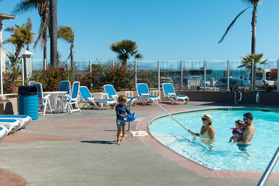 Rooftop pool deck with families swimming, lounge chairs, and ocean views behind glass railing