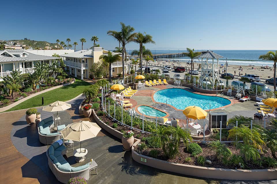 Beachfront resort with curved swimming pool, yellow umbrellas, ocean view, and beach pier in background