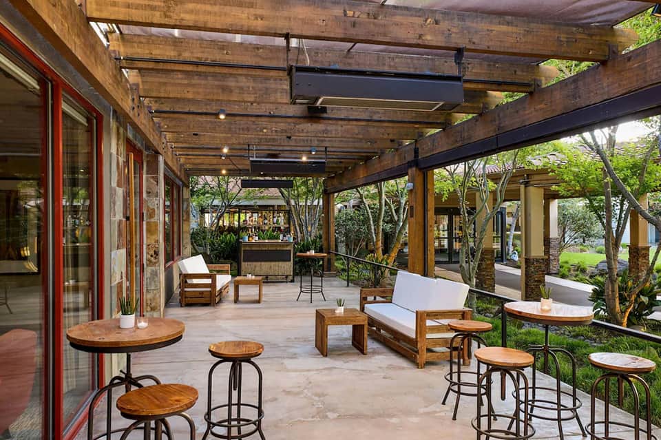 Covered patio bar area with wooden beams, tables, bar counter, and surrounding trees and greenery