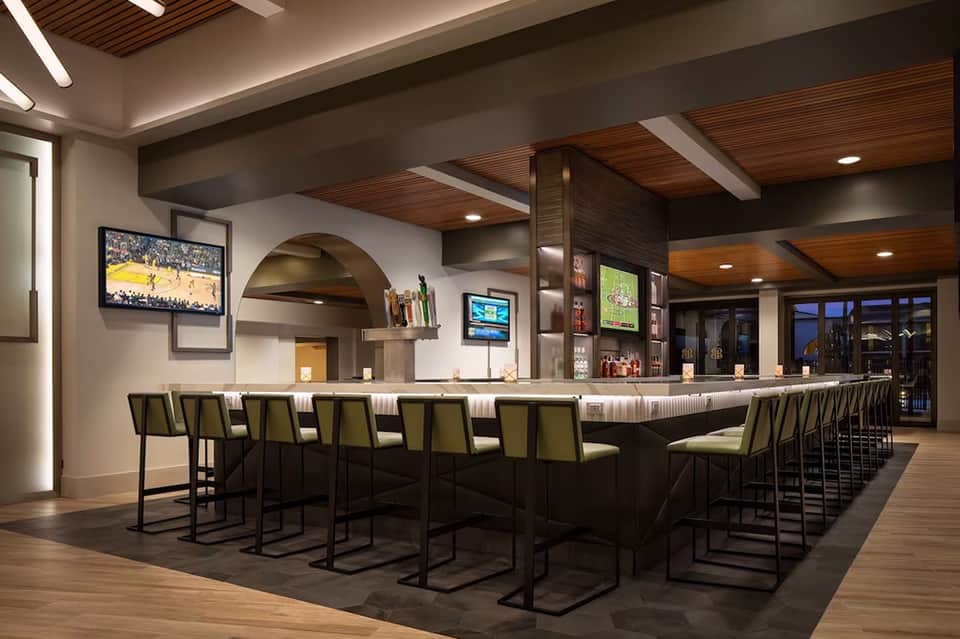 Modern hotel bar with dark wood ceiling beams, olive-green bar stools, multiple TV screens, and contemporary lighting