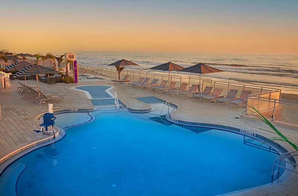 Beachfront pool deck at sunset with blue water, lounge chairs, umbrellas, and ocean view