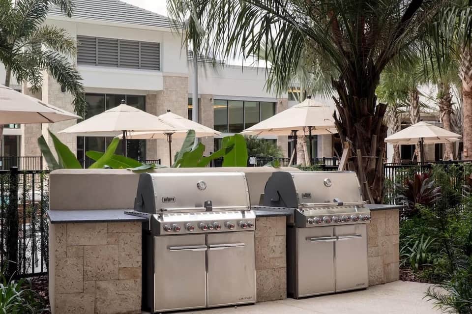Outdoor grilling station with two stainless steel Weber grills under umbrellas surrounded by palms