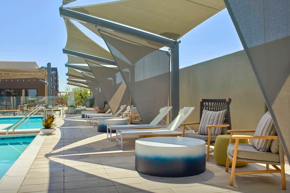 Outdoor pool area with lounge chairs under retractable shade canopies, fire pit, and city views