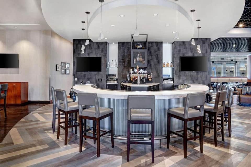 Modern hotel bar with curved white counter, dark gray tile wall, pendant lighting, and bar seating