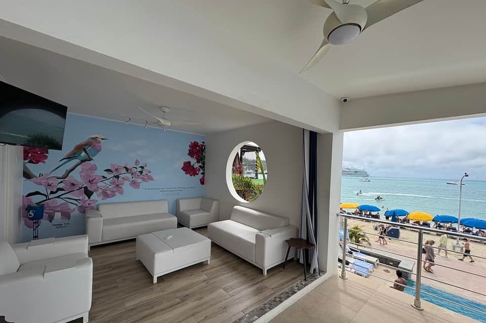 Modern oceanfront lounge with white furniture, large circular window, and beach view with blue umbrellas