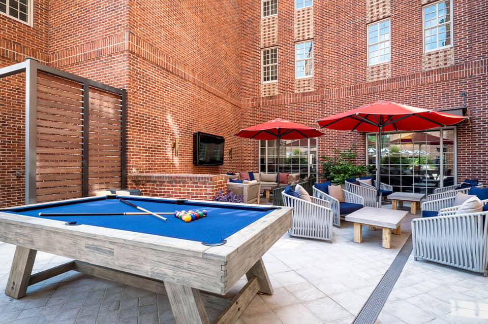 Courtyard patio with blue pool table, red umbrellas, white chairs, and brick building backdrop
