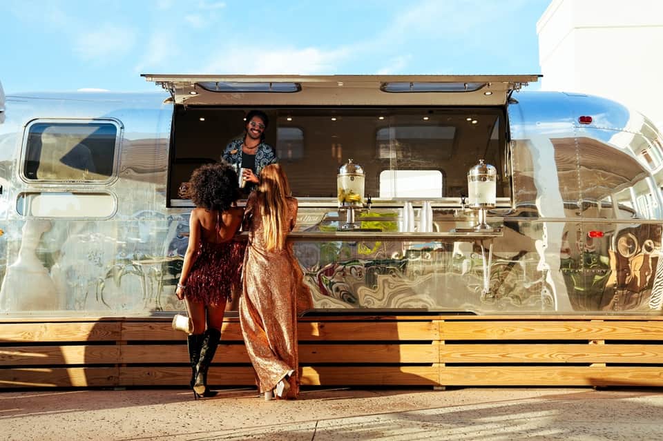 Women at vintage silver Airstream food truck service window on sunny day with beverages displayed