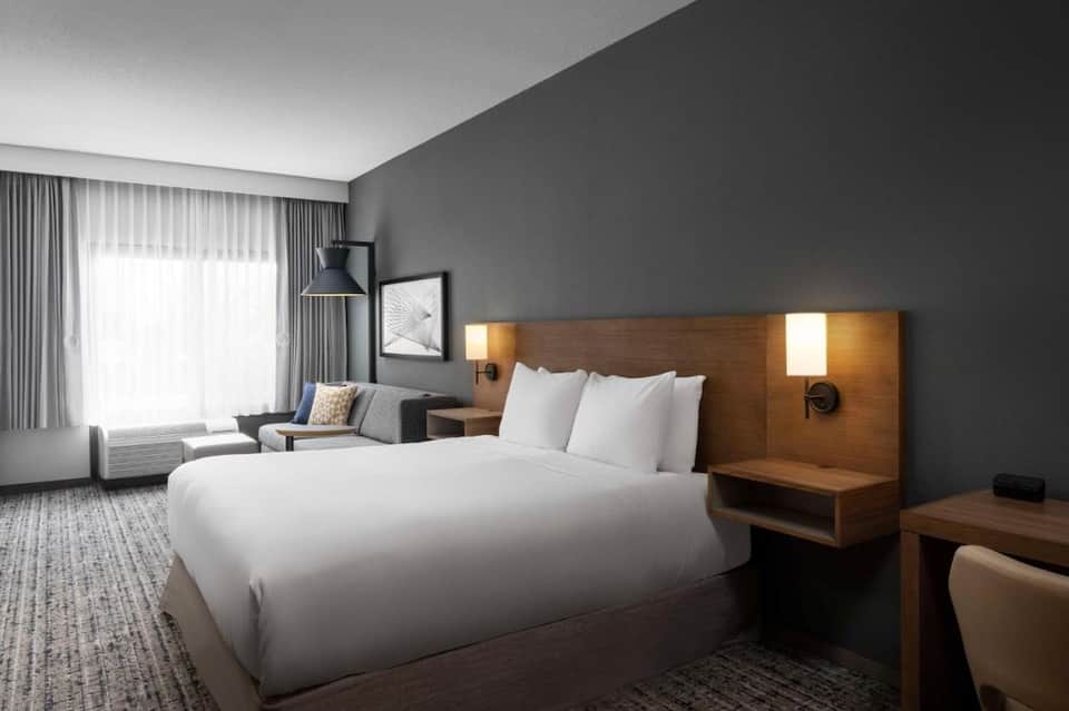 Contemporary hotel room with gray walls, wooden headboard with integrated lighting, white bedding, and seating area