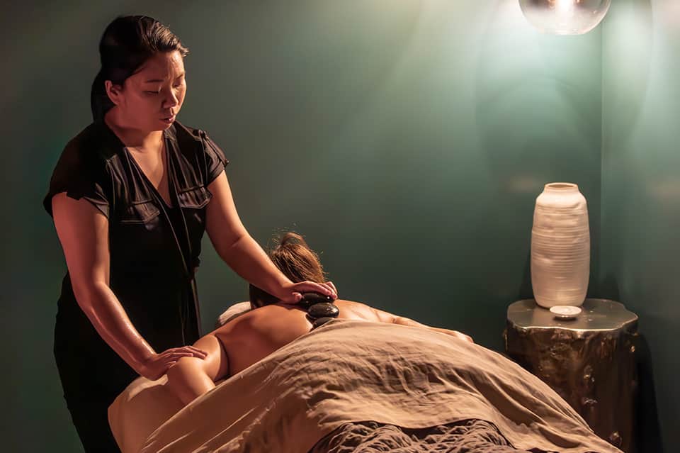 Spa therapist performing hot stone massage on client in peaceful green-walled treatment room with ambient lighting
