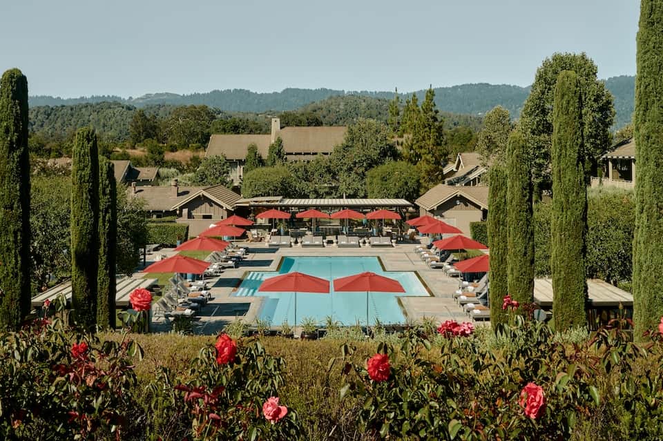 Resort pool surrounded by red umbrellas and cypress trees with forested hillside views in the background.