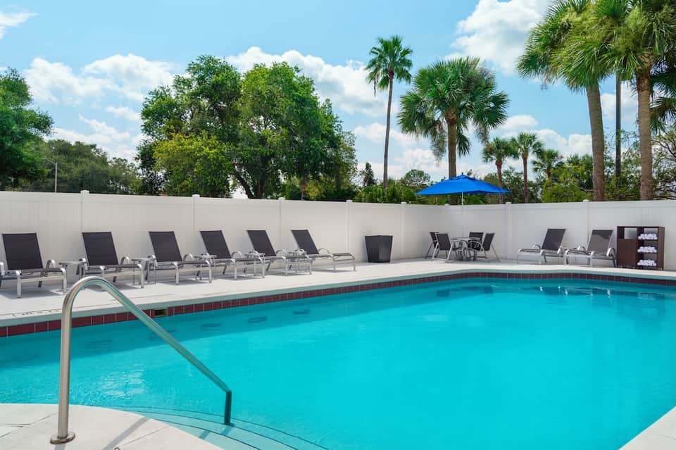 Resort pool with turquoise water, lounge chairs, white umbrella, and white privacy wall with palm trees in background.